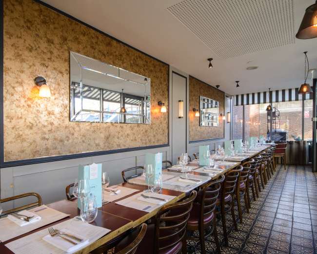 The image shows a dining area of a restaurant with long tables set for guests, featuring floral wallpaper, a large mirror, and pendant lighting.