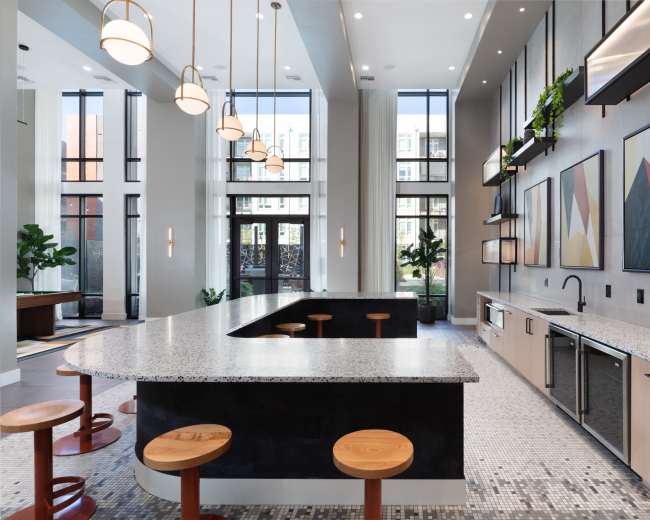 The image depicts a modern kitchen area featuring a large granite countertop with wooden stools, large windows allowing natural light, and geometric wall art.