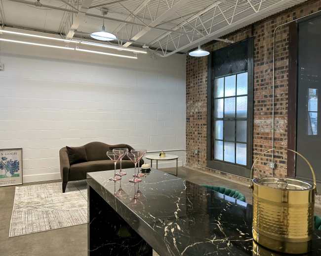 The image shows a modern office or lounge space with a dark sofa, a glass coffee table, a patterned rug, and a bar cart with glasses and a decorative item.