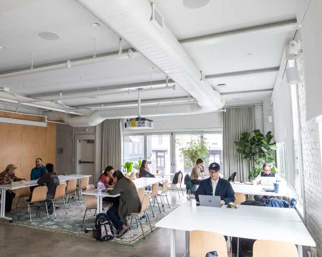 The image shows a modern coworking space with several people working at tables, large windows providing natural light, and plants adding greenery to the environment.