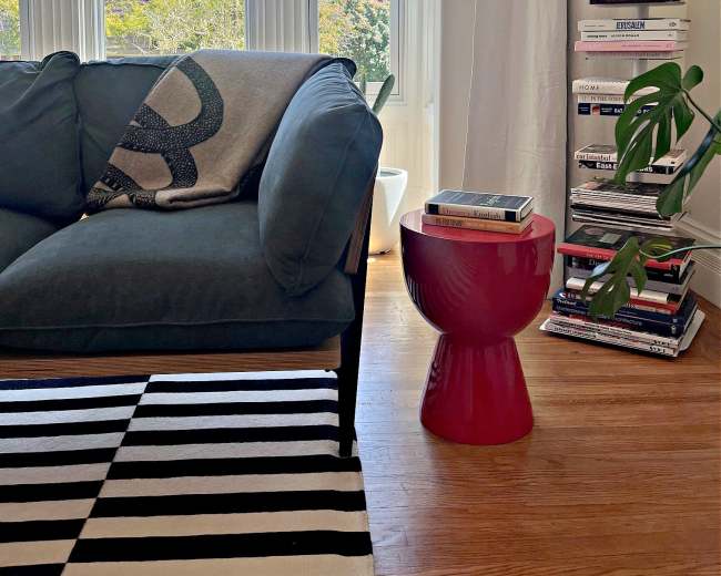 A living room features a dark green sofa, a striped rug, a red side table, and a stack of books beside a window with a view of trees.
