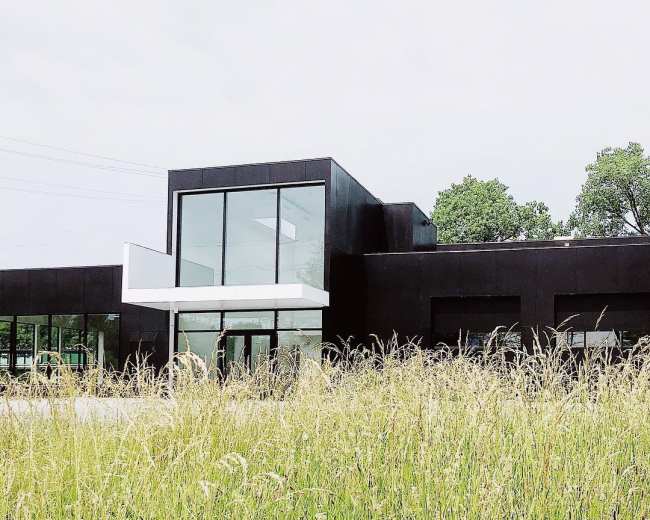 A modern black building with large glass windows is set against a backdrop of tall grass and trees under a cloudy sky.
