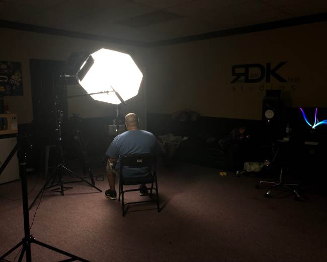 A man sits in a chair facing a large softbox light in a dimly lit recording studio.