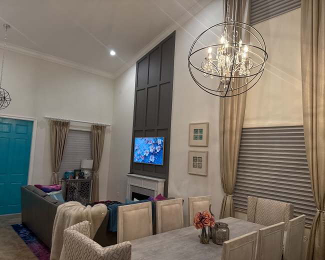 A modern living and dining area features a turquoise front door, a large wall-mounted television, a stylish chandelier, and a dining table surrounded by upholstered chairs.