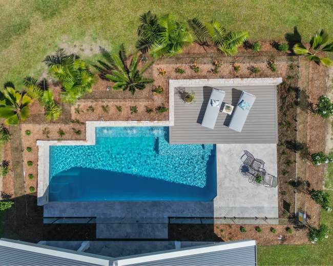 An aerial view shows a rectangular swimming pool surrounded by a landscaped area with palm trees and lounge chairs.