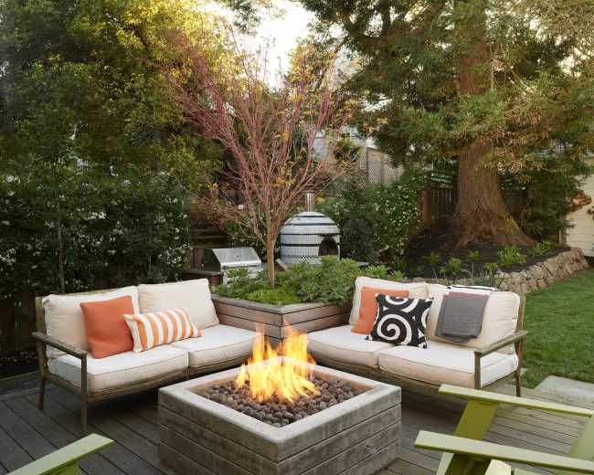 A cozy outdoor seating area features a fire pit surrounded by comfortable couches and vibrant green chairs, set against a backdrop of trees and garden greenery.