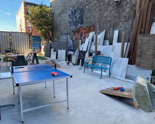 The image shows an outdoor area with a blue ping pong table, various construction materials, and scattered furniture against a graffiti-covered wall.