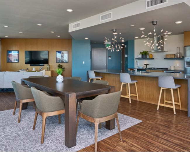 The image shows a modern open-concept living and dining area with a large dining table, stylish chairs, and a kitchen featuring sleek appliances and decorative lighting fixtures.