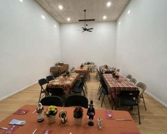 A large dining room features several tables set with checkered tablecloths and chairs arranged for a gathering, illuminated by natural and overhead lighting.