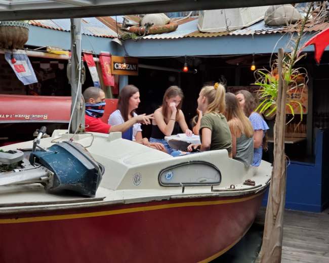 A group of young people is seated around a table on a boat, engaged in conversation at a casual waterfront venue.