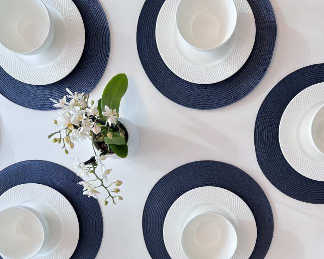 A neatly arranged dining table features six white bowls on textured navy blue placemats, accompanied by a small vase of white flowers.