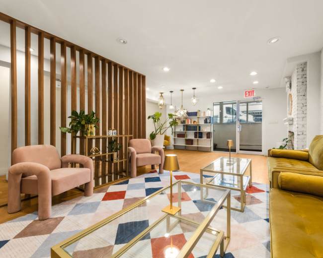 The image shows a modern lounge area featuring two pink armchairs, a yellow couch, a patterned rug, and a glass coffee table, with wooden slats and a bookshelf in the background.