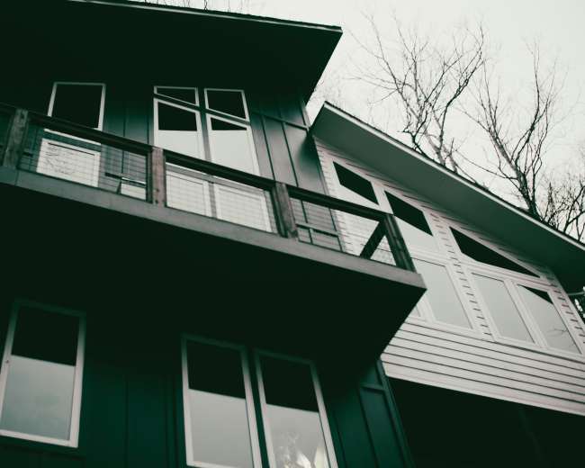 The image shows a modern house with multiple large windows and a balcony, set against a backdrop of bare trees.