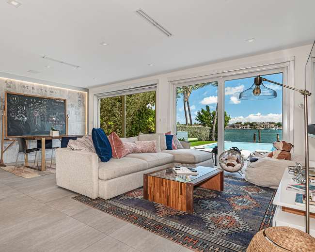 A modern living room features a large sofa, a coffee table, a chalkboard, and large windows overlooking a waterfront view with palm trees.