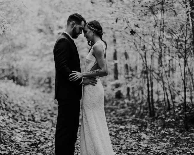 A couple stands close together in a forest, surrounded by trees and foliage.