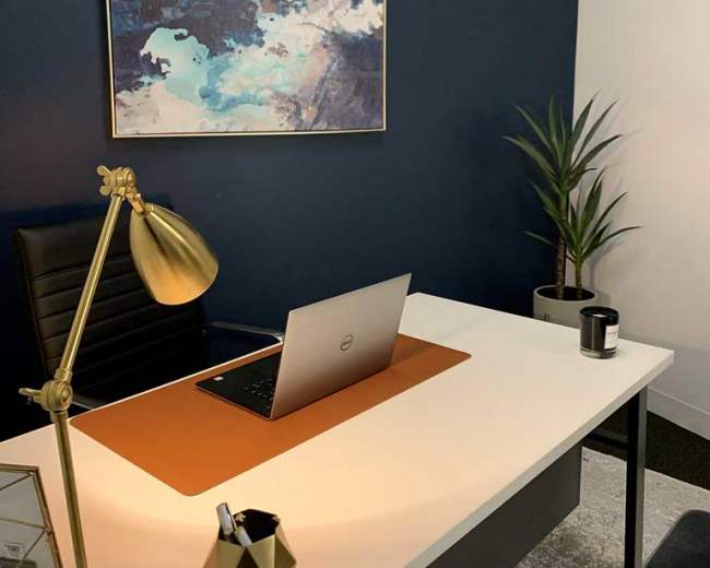 A modern office space features a white desk with a laptop, a golden lamp, a potted plant, and a blue accent wall adorned with abstract art.