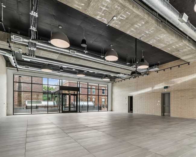 The image shows a spacious interior room with a polished tile floor, high ceilings, and large windows that allow natural light to enter.