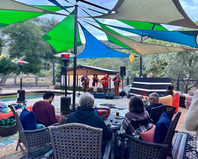 A small crowd sits in front of a live music performance under colorful canopies by a poolside area.