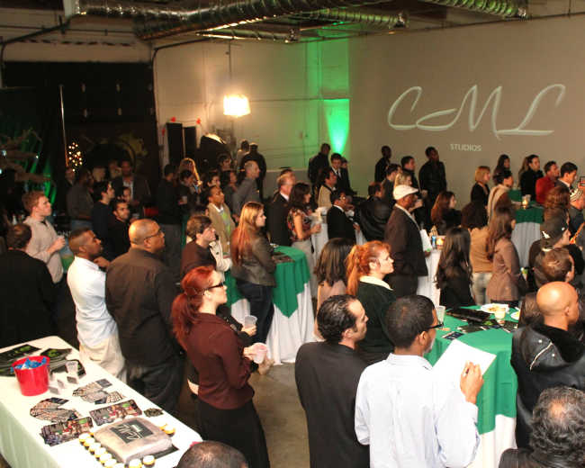 A large group of people stands in a spacious venue, some clapping and others engaged in conversation, with green and white tablecloths set up around the room.