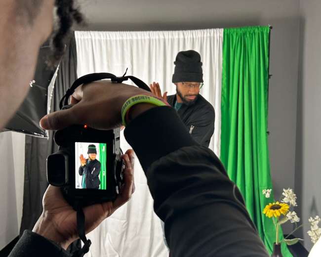 A person is photographing another individual posing in front of a green and white backdrop.