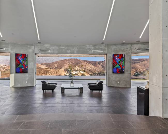 The image shows a modern interior space with concrete walls, large windows offering a view of mountains, and two black chairs facing a central coffee table.