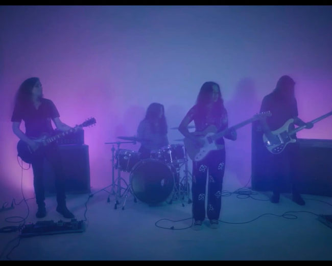 A band performs on stage with purple lighting, featuring two guitarists, a drummer, and a bassist.