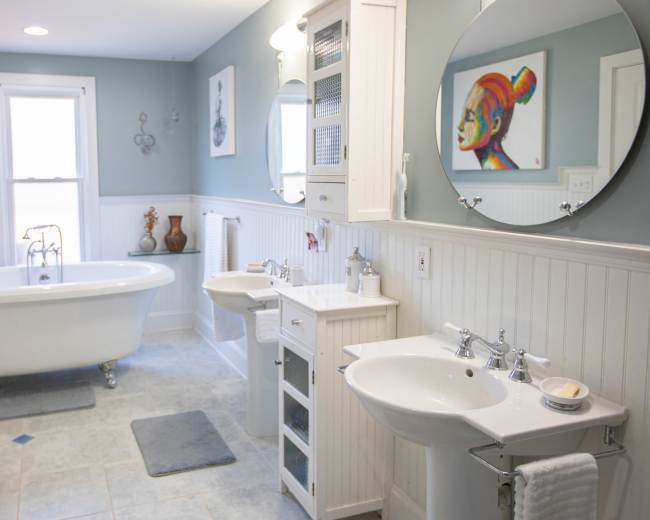 The image shows a bathroom featuring a freestanding tub, two sinks, and decorative artwork on the walls.