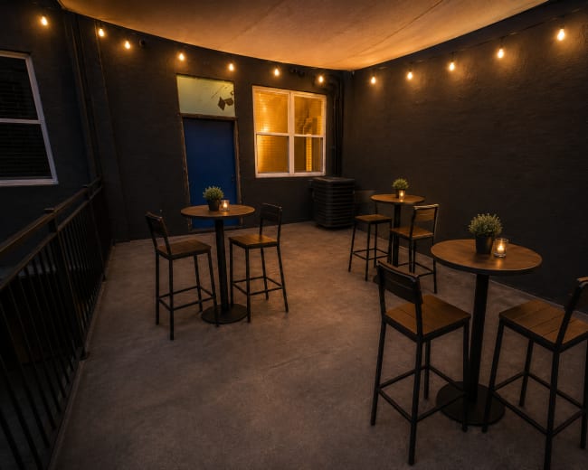 A dimly lit outdoor patio features several tall tables with chairs and small potted plants, creating an intimate setting.