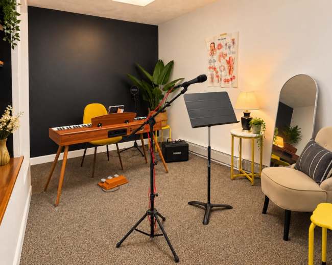 A small music practice room features a keyboard on a stand, a microphone on a stand, a music stand, and a chair, surrounded by plants and decorative elements.
