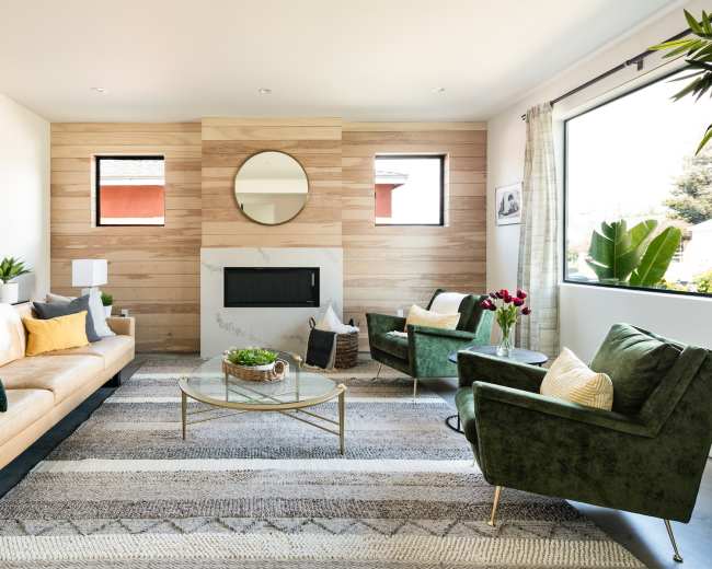 A modern living room features a beige sofa, green armchairs, a round mirror above a fireplace, and large windows letting in natural light.