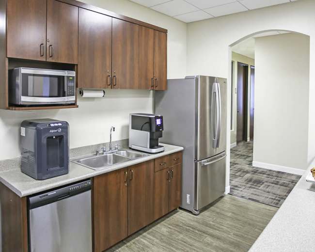 The image shows a modern kitchen area with wooden cabinetry, a sink, a microwave, a coffee machine, and a refrigerator, alongside a countertop displaying baked goods.