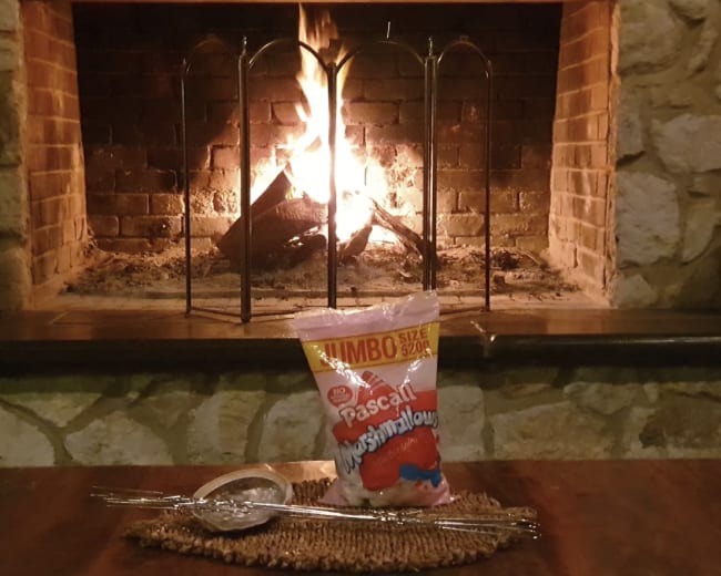A bag of jumbo marshmallows is placed on a table in front of a stone fireplace with flames burning.