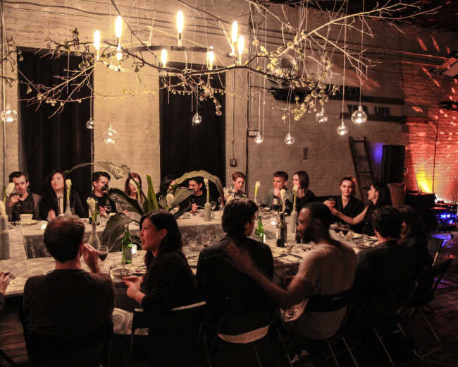 A group of people are seated around a long table in a dimly lit room, adorned with hanging lights and decorative branches, during what appears to be a dinner gathering.