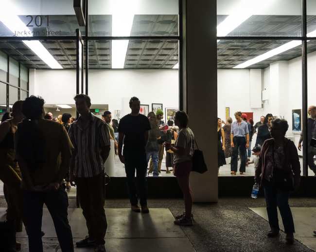 A crowd of people gathers outside an art gallery at night, with a well-lit exhibition visible through large glass windows.