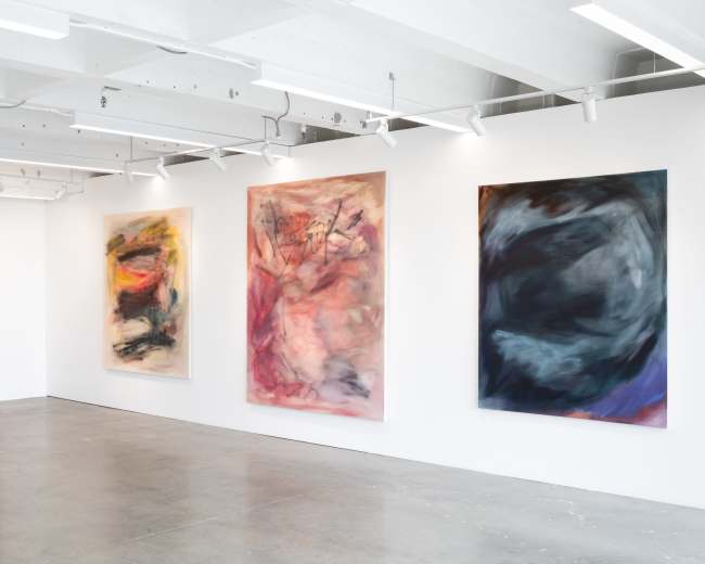 The image shows an art gallery with four large abstract paintings displayed on white walls in a spacious, brightly lit room.