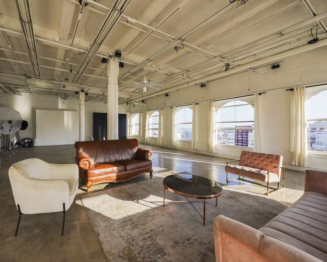 A spacious, well-lit studio featuring a mix of furniture, including a brown leather sofa, two chairs, and a coffee table, with large windows providing natural light.