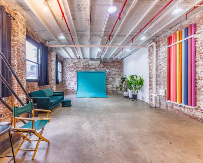 The image shows a spacious, industrial-style room with exposed brick walls, concrete flooring, a turquoise backdrop, and seating options including a green sofa and director's chairs.