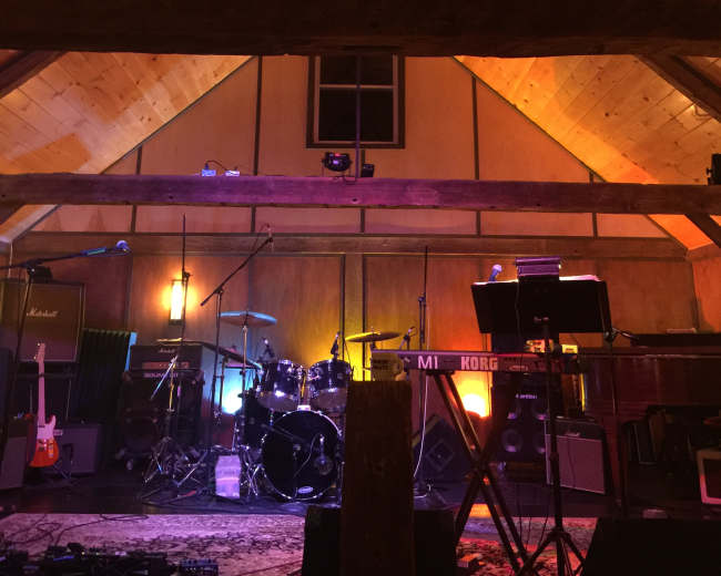A music performance space with wooden beams, stage lighting, and various musical instruments set up, including drums and keyboards.