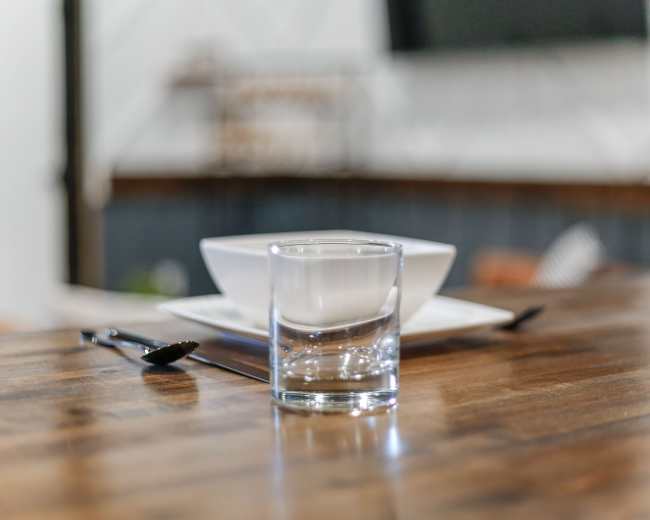 A clear glass filled with water sits on a wooden table next to a white bowl and a black spoon.