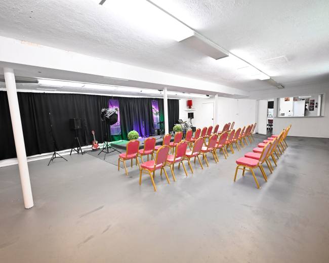 A spacious room with rows of pink chairs facing a black curtain, with a small stage setup at the front including microphones and lights.