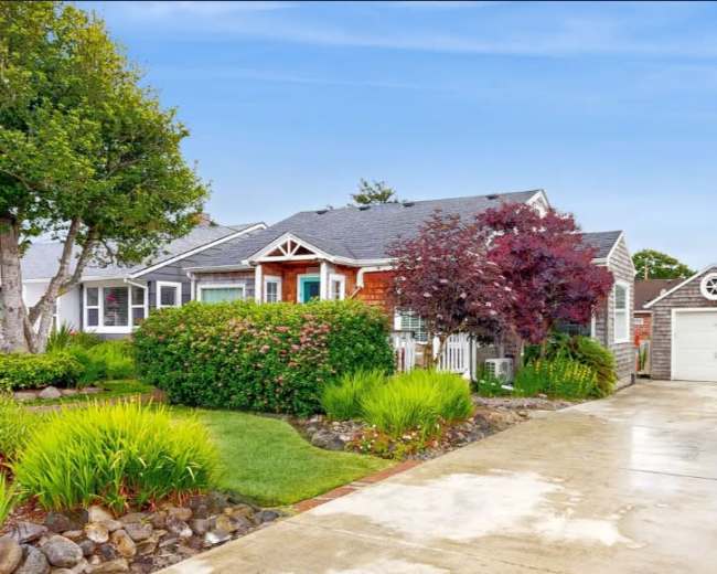 A single-story house with a well-maintained lawn and garden, accompanied by a garage on the side, is set against a clear blue sky.