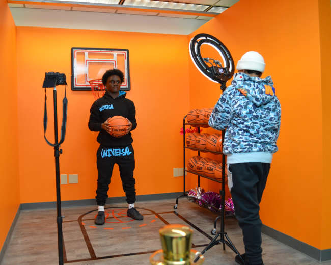 A person stands holding a basketball in front of a small basketball hoop set against an orange wall, while another person adjusts a camera on a tripod.