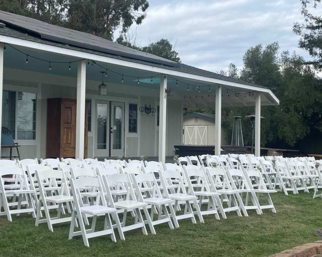 White folding chairs are arranged in neat rows facing a porch under a cloudy sky.