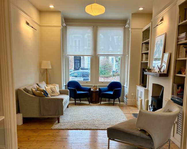 The image shows a cozy living room featuring two blue chairs, a small wooden coffee table, and a light-colored rug, with large windows allowing natural light to enter.