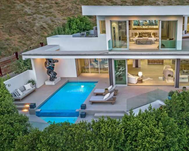 A modern house featuring multiple glass walls, a swimming pool, and a patio area surrounded by greenery.