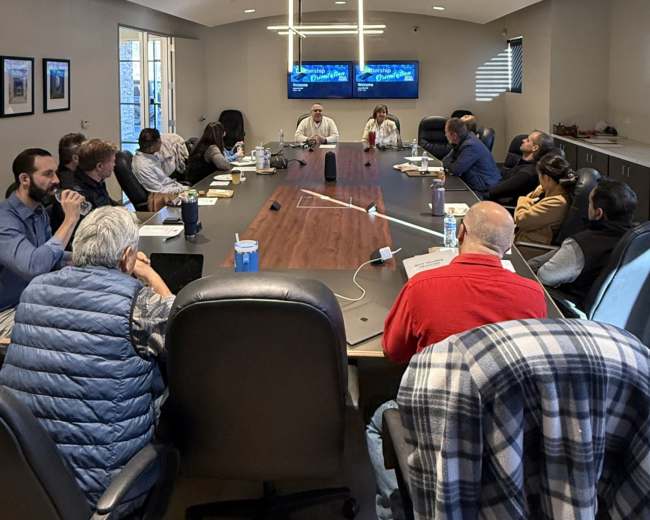 A group of professional individuals is seated around a large conference table in a well-lit meeting room, with multiple screens displaying information in the background.