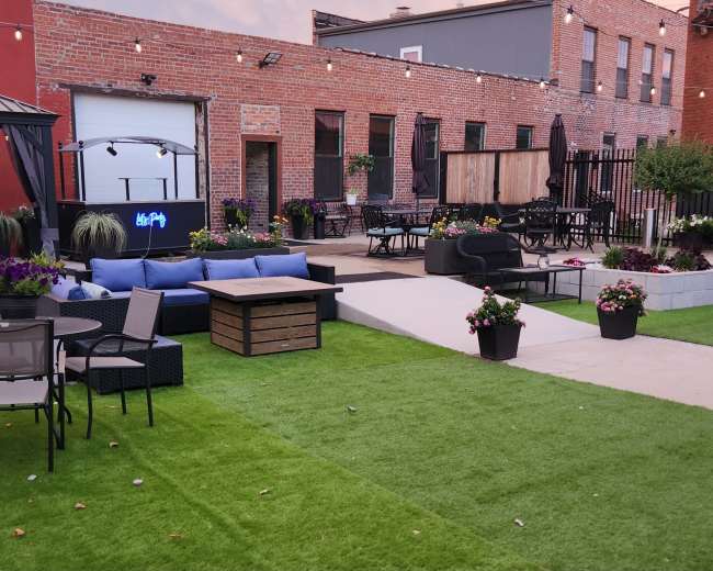 The image shows a landscaped outdoor patio area featuring artificial grass, a seating arrangement with sofas and chairs, potted plants, and string lights overhead.