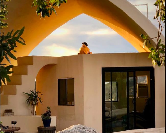 A person sits on the edge of a roof beneath a curved architectural structure during sunset.