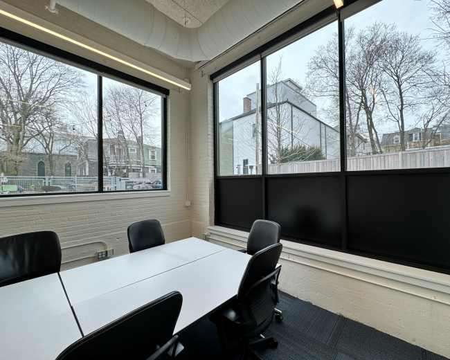A meeting room features a white table surrounded by black chairs, with large windows revealing a view of an outdoor area and neighboring buildings.