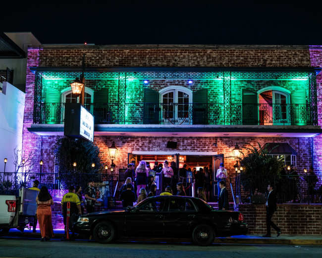 A lively gathering of people takes place outside a brightly lit, two-story brick building at night.